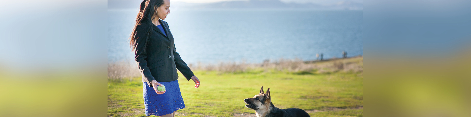 This photo is of Haben standing in a royal blue dress with a black blazer and with a light-green tennis ball in her hand. Her seeing-eye dog sees the ball and anticipates she will play by throwing it. It is a sunny day. They are standing on grass that is green with brown dirt patches. In the background, a body of water is blurred in the top third of the photo. 